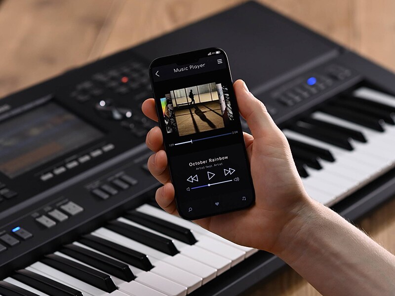 Clavier arrangeur Yamaha PSR-SX920 et une main tenant un smartphone affichant un écran de lecteur de musique
