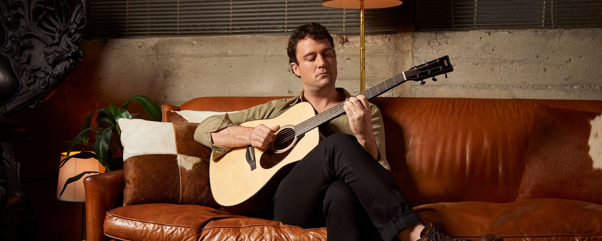 Guitarist Jordan Tice sits on a dark brown leather couch in a room lit with relaxing evening light playing an FG9 acoustic guitar.