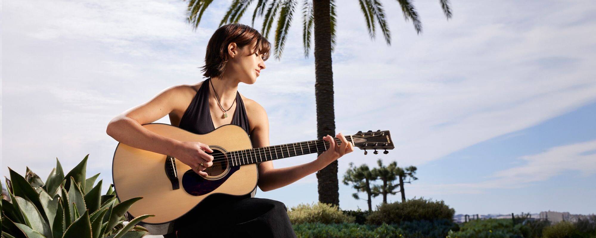 Guitarist Sophia James playing an FS9 guitar sits in front of a palm tree outside and plays an FS9 acoustic guitar.