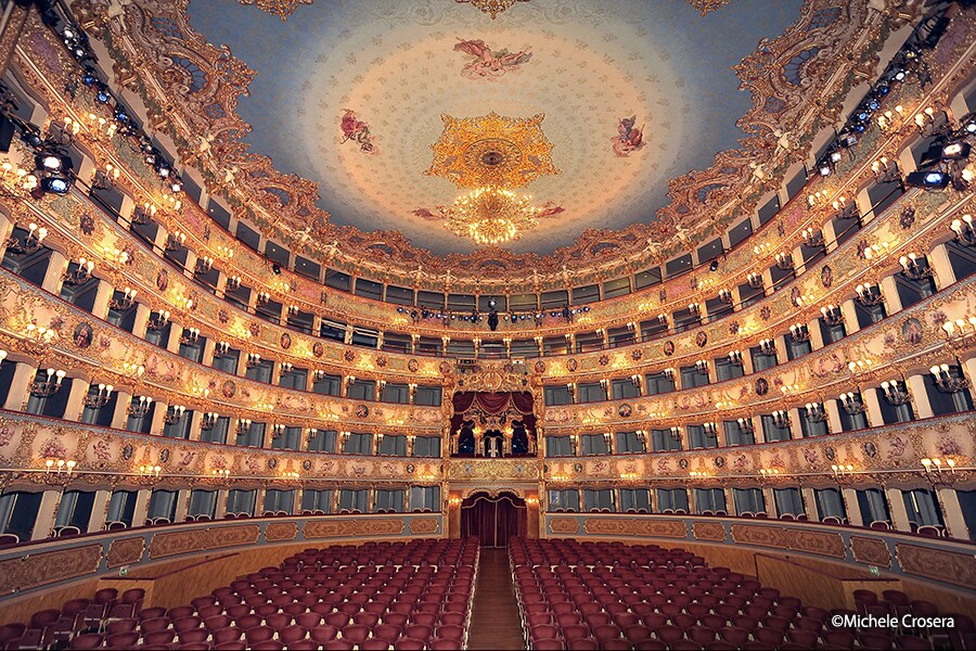 Wide interior view of Teatro La Fenice auditorium, highlighting the decorated ceiling, multiple balcony tiers, and red seating.